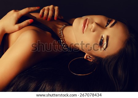 Fashion portrait of sexy tanned Asian lady with dark long hair bright evening makeup, wearing black lingerie and beautiful gold jewelry lying and posing at studio