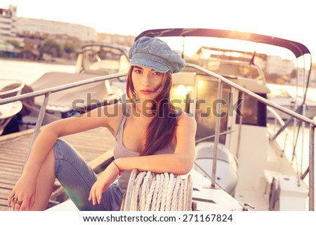 fashion outdoor photo of sexy sailor brunette girl wearing blue jeans,top and cap sitting and relaxing on a boat in the bay