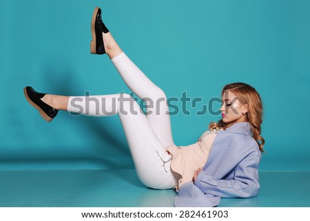 Fashion photo of sexy beautiful woman with blond curly hair, bright makeup wearing a blue coat,white trousers,black shoes lying on the floor at studio