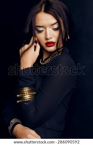 Fashion photo of sexy Indian woman with dark hair and bright make-up wearing black shirt and  luxurious gold necklace,bracelets, earrings posing at studio