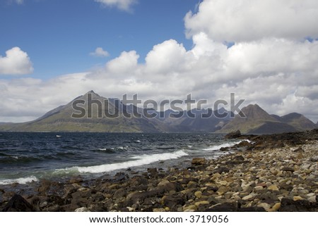 Sunlit Cullin Mountains from Elgol on the Isle of Skye. Waves crash on to a rocky beach in the foreground