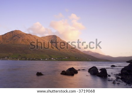 Sunset over distant mountains, fluffy white clouds are tinged with orange and frame the mountain slopes. Calm water and silhouetted rocks form the foreground