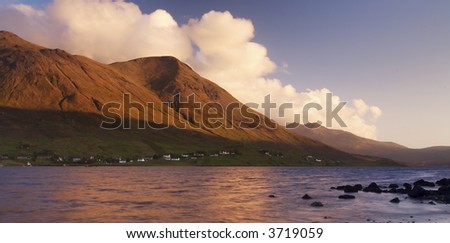 Sunset over distant mountains, fluffy white clouds are tinged with orange and frame the mountain slopes. Calm water and silhouetted rocks form the foreground