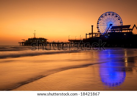 The Neon lit Ferris Wheel of Santa Monica Pier is reflected in the wet sand of the beach at Sunset.