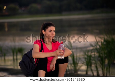 Young woman athlete resting after a long run in the evening. She is sitting near a lake with a bottle of water in the hand.