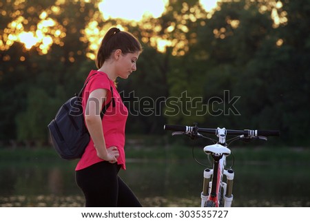 Young woman athlete with her mountain bicycle in the evening. She is stopped for resting near a lake with sunset behind.