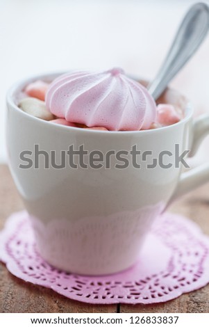 Closeup of cup of hot chocolate with pink meringue and spoon resting on small pink table mat on wooden table