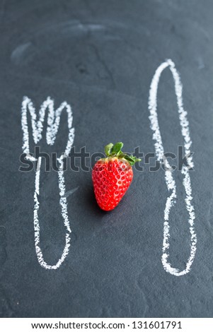 One fresh whole strawberry and chalk drawings of knife and fork on dark background