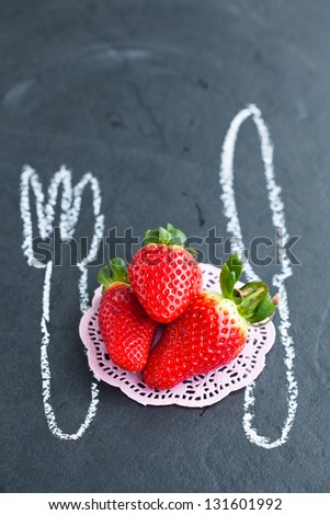 Three fresh whole strawberries and chalk drawings of knife and fork on dark background