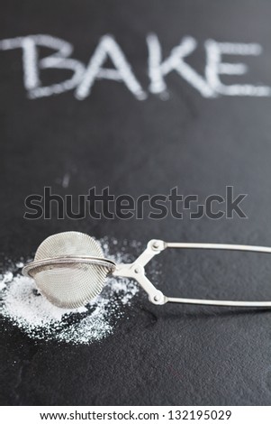 Closeup of sieve and icing sugar with word bake written in chalk on dark textured background