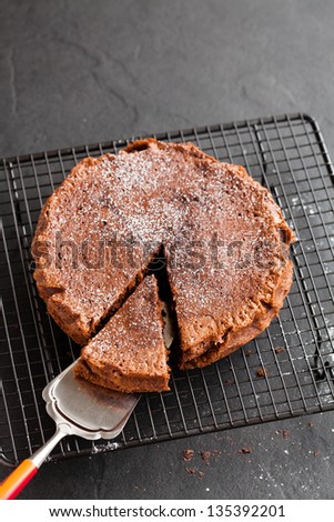 Freshly baked homemade chocolate cake on metal cooling rack with cut slice and cake server utensil on dark background