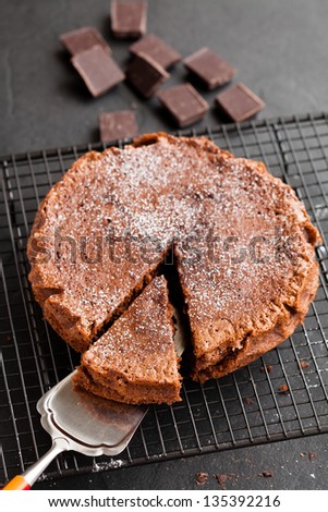 Freshly baked homemade chocolate cake on metal cooling rack with cut slice and cake server utensil and chocolate squares on dark background