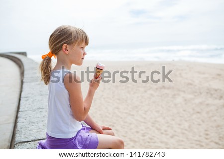 Young cute girl holding ice cream while sitting on wall at beach