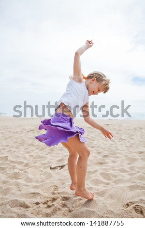 Happy barefoot young girl in purple skirt and white top jumping in sand on beach