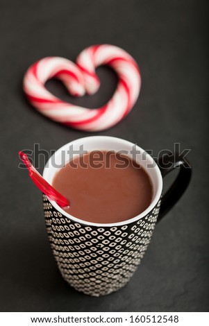 Closeup of mug of hot chocolate with red plastic spoon and hard candy heart with red swirls in background