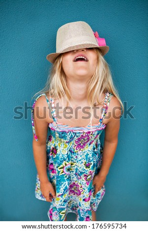 Portrait of stylish cute young girl against blue wall background with hat covering eyes and funny expression