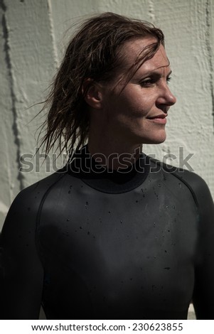 Attractive brunette female swimmer wearing black wetsuit with wet hair and looking calmly with slight smile and sunlight in face