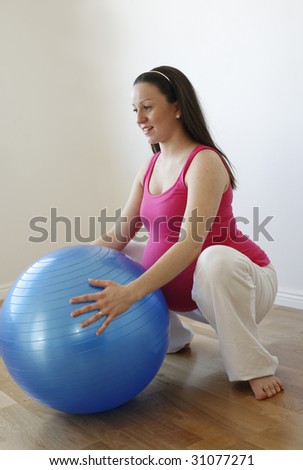 A young smiling pregnant woman in a pink shirt doing a squatting exercise with a blue fitness ball while standing up.
