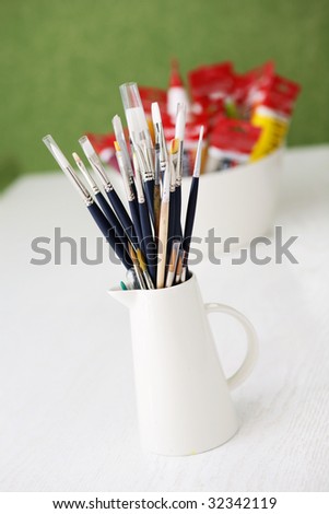 A set of paintbrushes in a jug and tubes of paint in a bowl on a white table in an outdoor setting with green grass in the background.