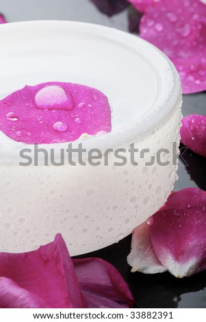 A close-up of a health and beauty spa decoration with a pink flower petal and water droplet resting on the water surface in an opaque round bowl surrounded by more pink flower petals.