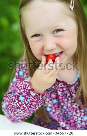 A close-up of a smiling little girl at the edge of a white table eating a delicious strawberry outdoors in summer with a green grass background.