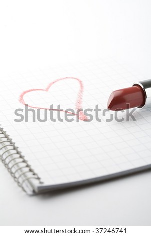 A close-up of a tube of red lipstick and a heart shape on a notepad with a spiral binding.