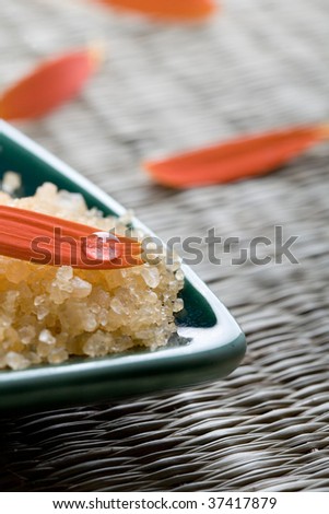 A close-up of an orange flower petal and water droplet resting on yellow bath salt crystals in a turquoise colored dish used in a health and beauty spa.