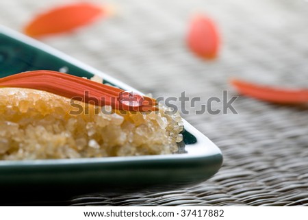 A close-up of an orange flower petal and water droplet resting on yellow bath salt crystals in a turquoise colored dish used in a health and beauty spa.
