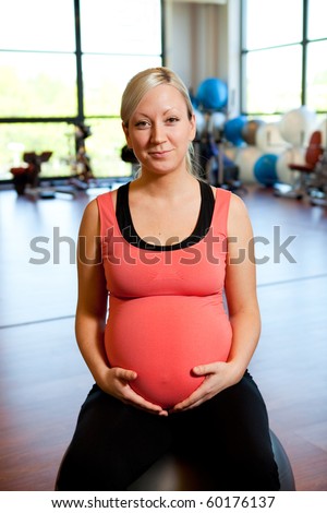 A smiling pregnant woman sitting on a fitness ball in a gym while relaxing and holding her belly.