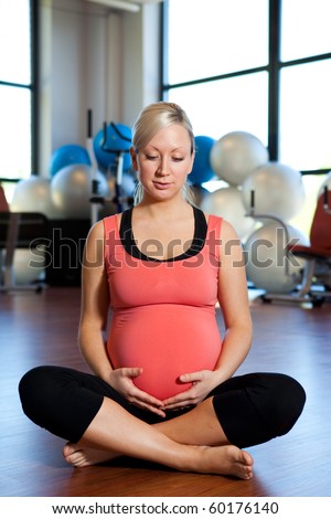 A pregnant woman sitting on the floor of a gym and holding her belly while relaxing.