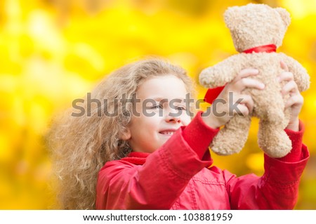Beautiful and happy little girl holding teddy bear with her hands. Smiling and looking at the toy. Yellow autumn trees in background.