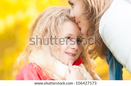 Beautiful and happy young mother kissing her little daughter on forehead. Both smiling. Autumn trees in background. Fall season.