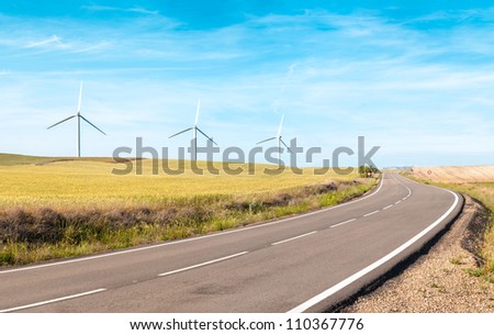 Wind turbine on green and yellow field. Empty road in foreground, blue sky with clouds in background. Alternative energy source, production and power generation. Ecology and freedom concept.