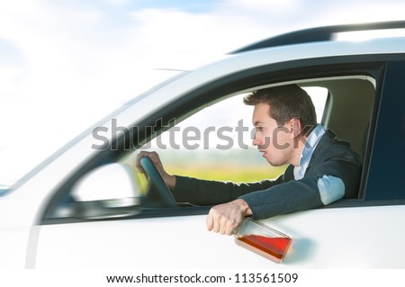 Drunk man holding bottle of whiskey in left hand. Person driving and drinking alcohol. Outdoor action with young guy inside white car. Risky travel. Bright and sunny summer day in background.