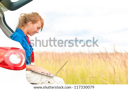 Young happy girl sitting and working on computer. Beautiful woman near car in meadow holding laptop. Person working outdoor. Tail light of car in foreground and field with sky in background.