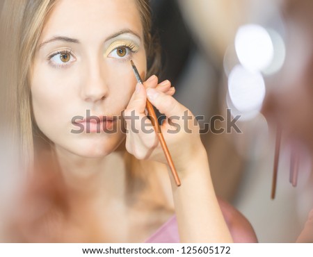 Beautiful young woman getting make-up done. Make up artist applying eye shadow with brush. Reflection in mirror with blurs in background. Cosmetics and make-up accessories.