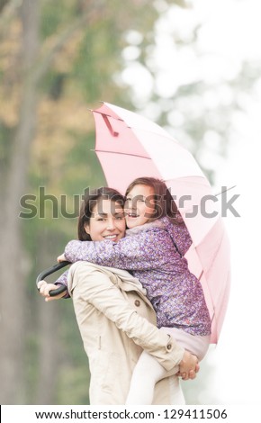 Beautiful mother embracing daughter under umbrella. Smiling woman and girl in rainy weather. Portrait of happy mom and kid. Young parent with child walking in park in autumn. Family outdoor activity.