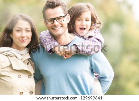 Young happy family of three having fun together outdoor. Pretty little daughter on her father back. Parents and girl look happy and smile. Happiness and harmony in family life. Family fun outside.