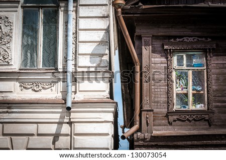 Two houses of different style in Russia. Old dark wooden house with carved window and white brick house with stucco work. Traditional Russian architecture in contrast.