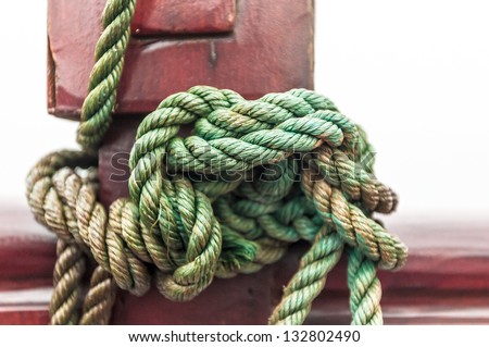 Marine rope tied into knot in foreground, white background. Close-up of nautical equipment for mooring. Focus on detail of ship. Green twisted rope wrapped around red wooden deck. Sea travel.
