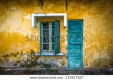 Outside view of deserted house with details in Vietnam. Old and grungy yellow wall with window and worn blue door. Abandoned place with lock on door, half-open sun blinds and metal grating on window.