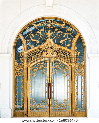 Beautiful Arched Doorway. Door Made Of Wood, Gold And Glass Reflecting Arch. White Wall Of Building With Elegant Gilded Entrance. Old Style Of Door With Golden Ornate Details. Tourist Attraction.