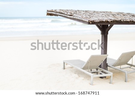 Two empty chaise longues under wooden shed on beach with blue ocean waves in background. Heaven and paradise atmosphere. Summer holidays and vacations in tropical countries.
