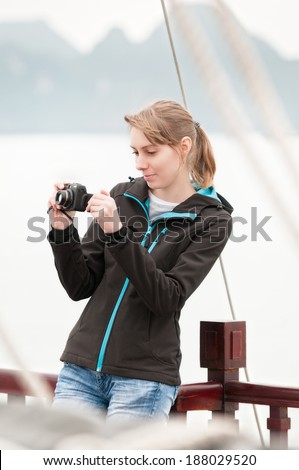 Woman standing on deck of ship at Halong Bay and holding camera. Pretty girl at cruise ship in Vietnam, Asia. Mountains and bay in background. Beautiful woman in jeans and jacket. Active lifestyle.