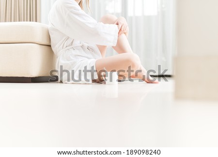 Woman in white bathrobe sitting on floor in hotel room. Beautiful and elegant interior. Bedroom in soft colors. Cup of coffee next to girl. Person relaxing and drinking tea.