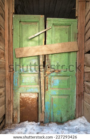 Skewed old green door in wooden house. Torn lock, clogged entrance. Dirty snow in from of building. Winter in city.