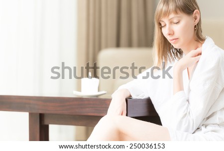 Pretty woman in white bathrobe relaxing at table in hotel room. Cup of coffee on table. Modern interior. Window and curtain in background. Beautiful girl relaxing and drinking tea in morning.