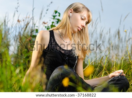 beautiful young girl sitting and listening music outside with sky and grass in background