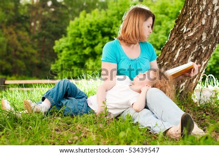 young mother sitting in grass under tree and reading book to her small daughter who is lying on knees of her mother and sleeping