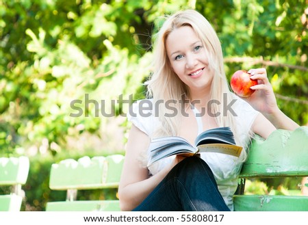 young woman sitting on bench in park, reading book, eating red apple, smiling and looking into the camera
